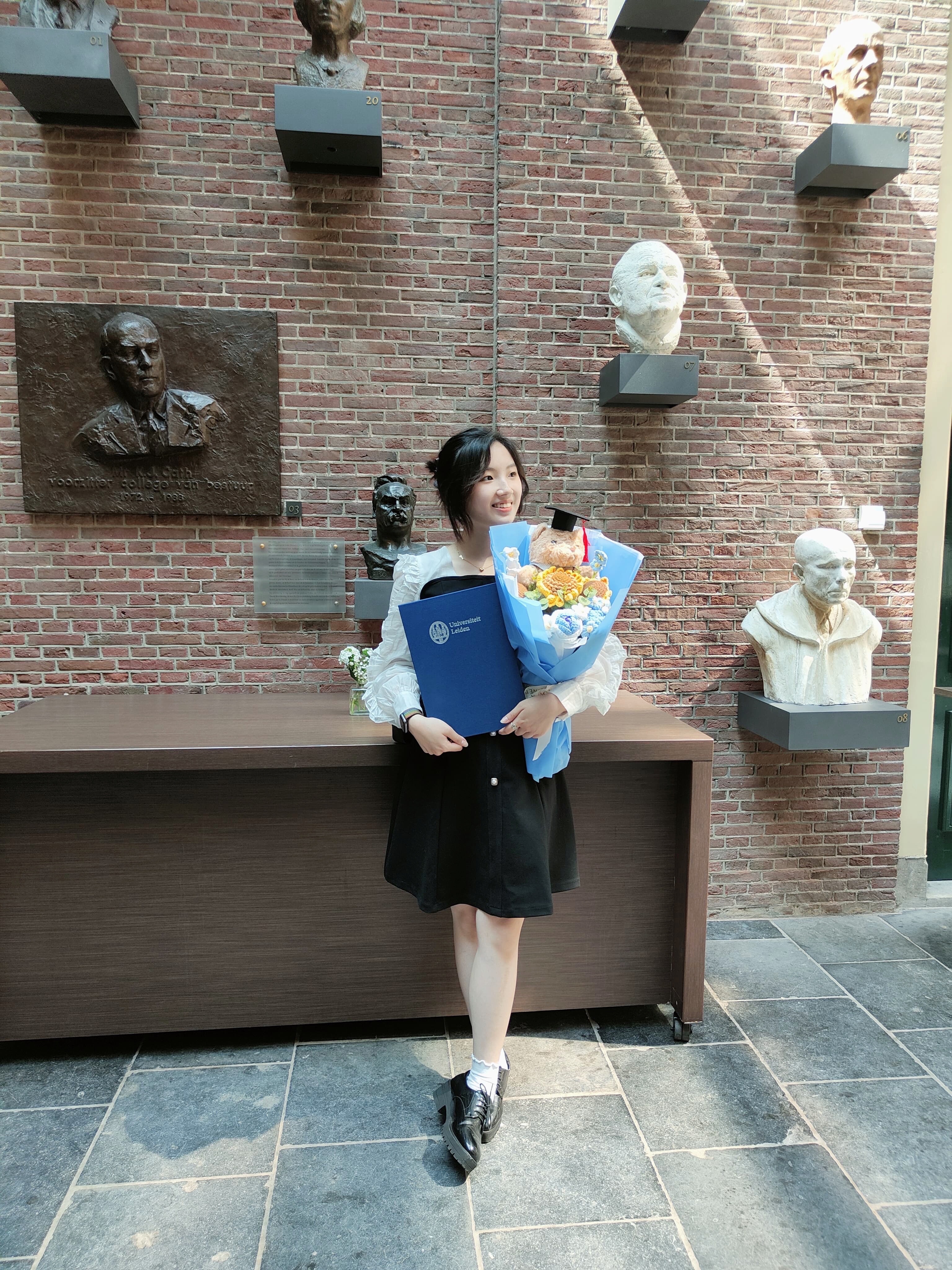 Chiini smiling in graduation gown, holding a diploma and flowers in front of a brick wall with sculpted busts